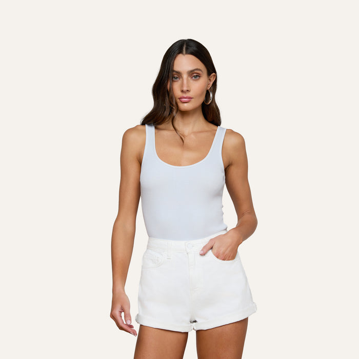 Woman with long brown hair wearing the Nica Scoopneck Bodysuit, styled with white shorts and standing against a plain light background with one hand in her pocket.