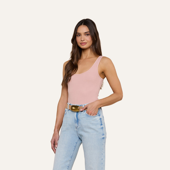 A woman with long brown hair stands and smiles against a plain, off-white background, wearing a light pink Nica Scoopneck Bodysuit with a sculpted fit and light blue high-waisted jeans accessorized with a belt.