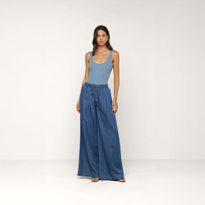 A woman stands against a plain background, wearing the Nica Scoopneck Bodysuit in light blue as a base layer and wide-leg blue denim pants with a drawstring waist. She has long hair and a neutral expression, with her arms relaxed at her sides.