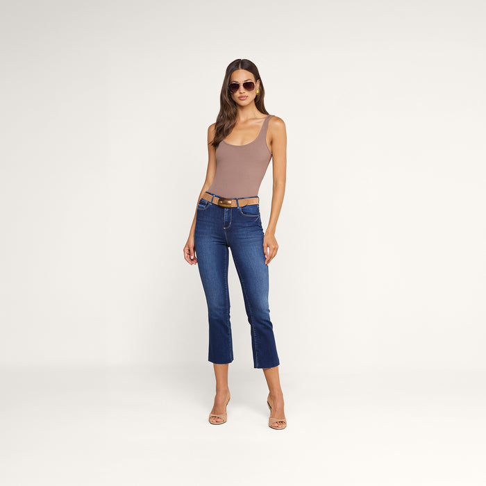 A woman wearing sunglasses, the Nica Scoopneck Bodysuit in taupe with a stretch rib and sculpted fit, blue cropped jeans, and nude heels stands against a plain white background.
