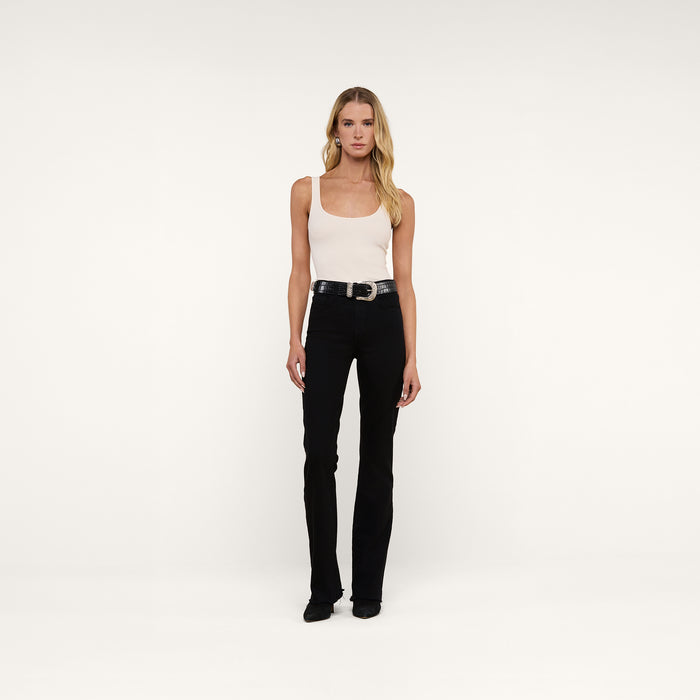 A person with long blonde hair wears the Nica Scoopneck Bodysuit— a sleeveless beige stretch-cotton top with a scoop neckline—paired with a black belt, black pants, and black shoes, standing against a plain white background.