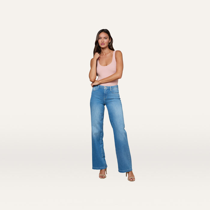 A woman stands posing in the Nica Scoopneck Bodysuit, a light pink high-stretch ribbed style, paired with blue high-waisted wide-leg jeans and high-heeled sandals against a plain white background.