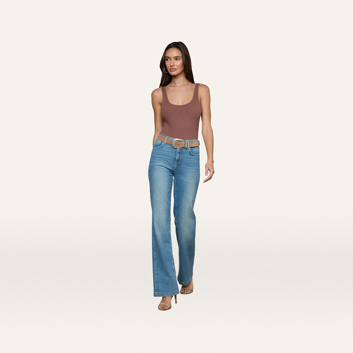 A woman wearing the Nica Scoopneck Bodysuit, styled with blue high-waisted jeans, a belt, and heels, walks against a plain white background.