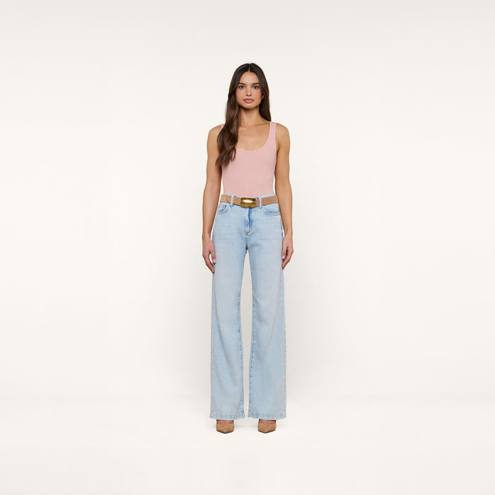 A woman with long brown hair and a neutral expression stands against a plain white background, wearing the Nica Scoopneck Bodysuit in light pink, paired with light blue high-waisted wide-leg jeans, a brown belt, and beige shoes.