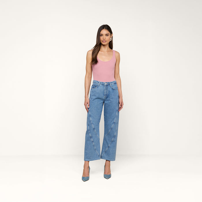A woman stands against a white background wearing the Nica Scoopneck Bodysuit in pink, paired with high-waisted blue jeans and blue heels. Her arms are relaxed by her sides, and she has a neutral expression.
