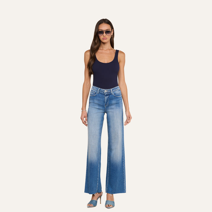 A woman wearing sunglasses, the Nica Scoopneck Bodysuit styled as a sculpted fit base layer, light blue flared jeans, and blue heels stands against a plain light background.