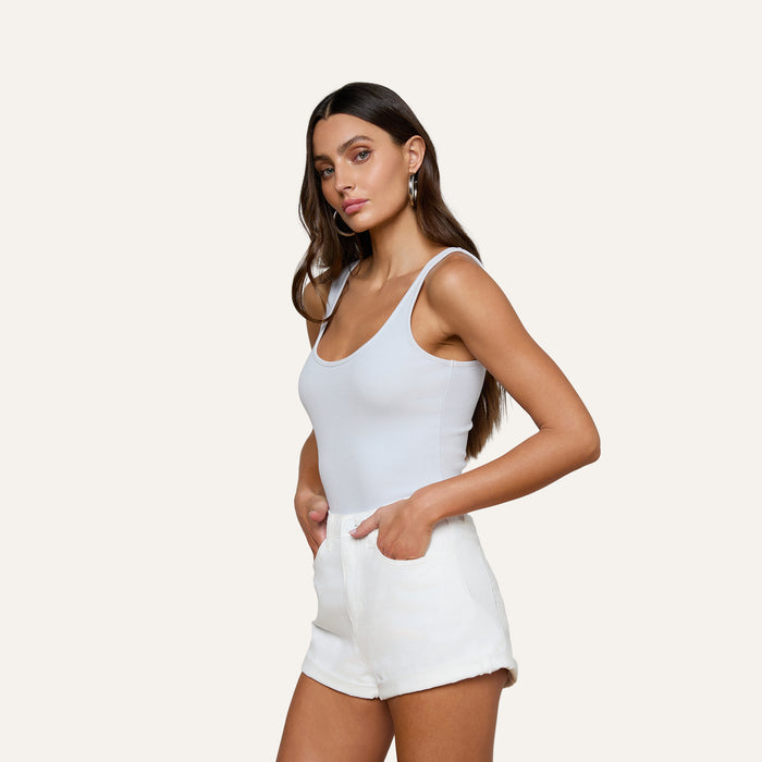 A woman with long brown hair wears the Nica Scoopneck Bodysuit and white shorts, standing against a plain light background with her hands in her shorts pockets, looking toward the camera.
