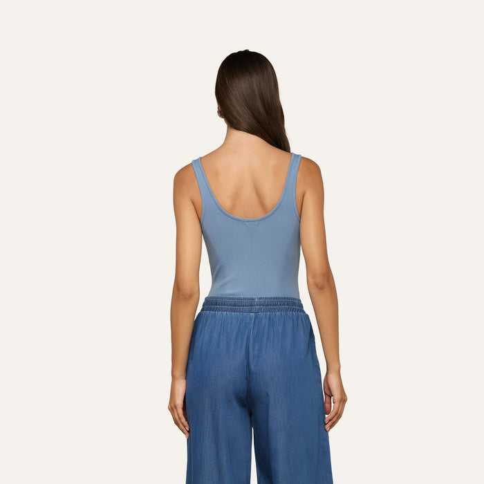 A woman with long brown hair is shown from the back, wearing the Nica Scoopneck Bodysuit in light blue with a sculpted fit and loose blue pants against a plain light background.