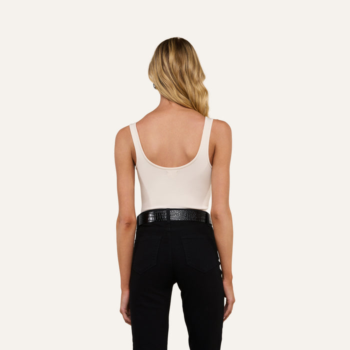 A woman with long blonde hair stands with her back to the camera against a light background, wearing the Nica Scoopneck Bodysuit—a sleeveless, light beige top with a scoop neckline—paired with black pants and a textured black belt. Crafted from stretch-cotton for added comfort, the bodysuit makes an ideal layering piece.