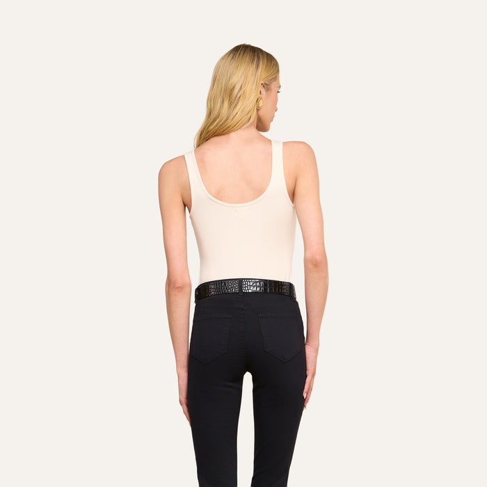 A woman with long blonde hair stands facing away, wearing a sleeveless beige Nica Scoopneck Bodysuit tucked into high-waisted black pants with a black textured belt, set against a plain light background.