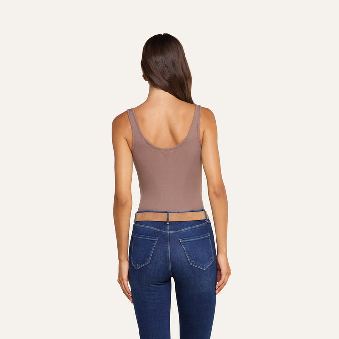 A woman with long brown hair is shown from the back, wearing the Nica Scoopneck Bodysuit in a sculpted, sleeveless fit paired with high-waisted blue jeans featuring a visible waistband. The background is plain and light-colored.