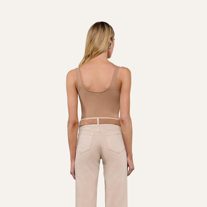 A woman with long blonde hair, dressed in a tan Nica Scoopneck Bodysuit as a stretch rib base layer and light beige pants, stands facing away from the camera against a plain light background.