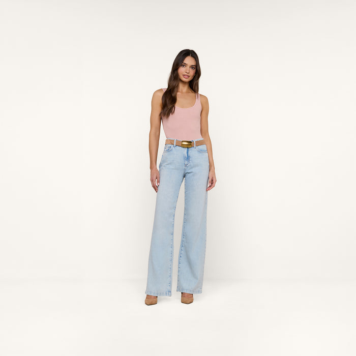 A woman stands against a plain white background, wearing a light pink Nica Scoopneck Bodysuit with a sculpted fit, light blue high-waisted wide-leg jeans, and beige high heels.