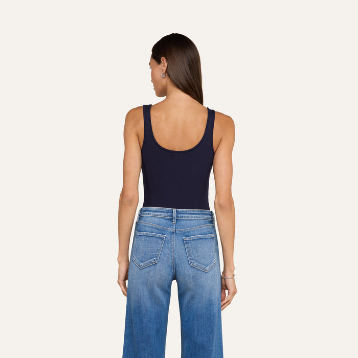 A woman with long brown hair, wearing the Nica Scoopneck Bodysuit in navy and high-waisted blue jeans, stands facing away, highlighting the sculpted fit of her outfit against a plain light background.