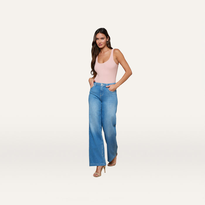 A woman wearing the Nica Scoopneck Bodysuit in light pink, paired with high-waisted blue wide-leg jeans and beige heels, stands against a plain white background, looking at the camera with her hands in her pockets.