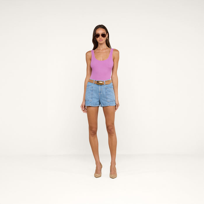 Woman wearing sunglasses, the Nica Scoopneck Bodysuit in pink with a sculpted fit, denim shorts with a belt, and beige heels stands against a plain white background.