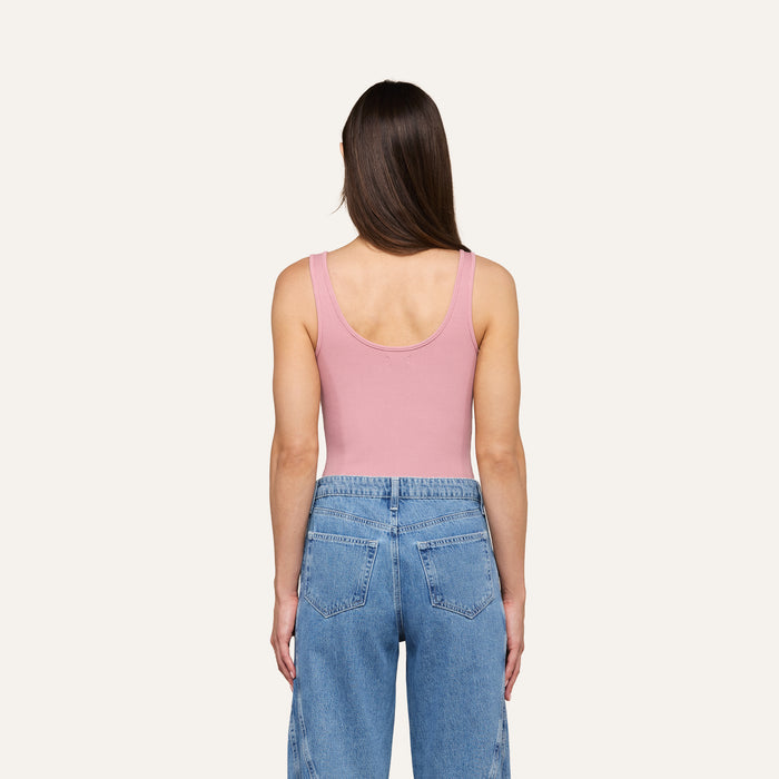 A woman with straight brown hair is standing with her back to the camera against an off-white background, wearing the Nica Scoopneck Bodysuit in pink and blue jeans.
