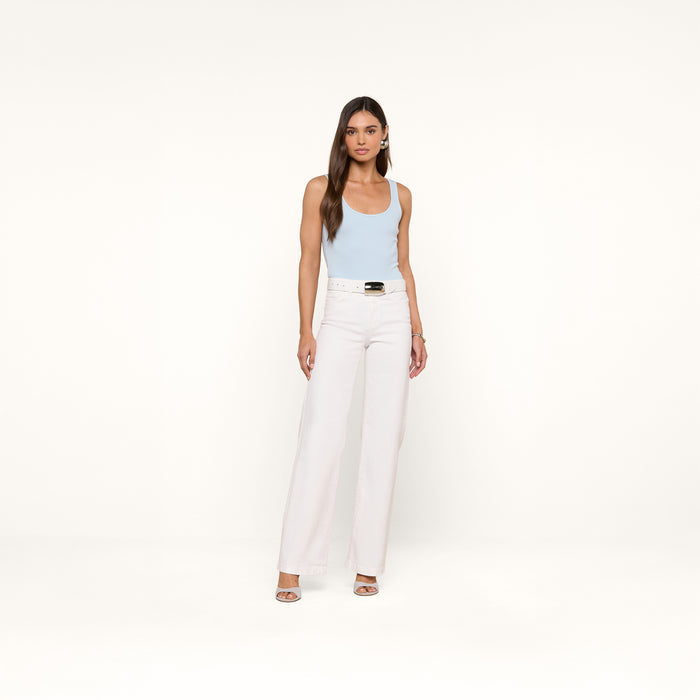 A woman with long brown hair and a neutral expression stands against a plain white background, wearing the Nica Scoopneck Bodysuit in light blue, paired with high-waisted white wide-leg pants, a white belt, and open-toe heels.
