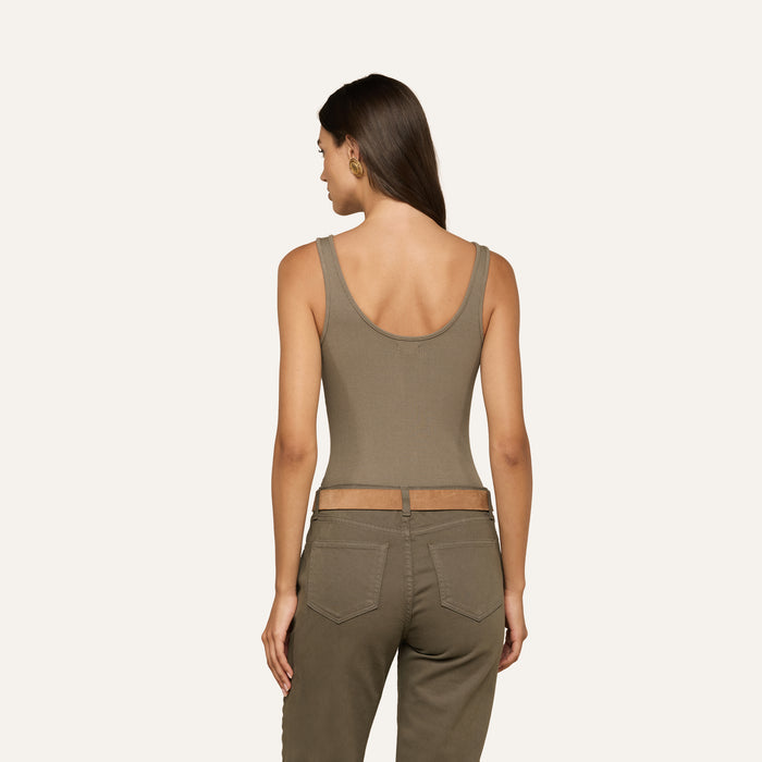 A woman stands facing away, wearing the Nica Scoopneck Bodysuit in olive—a sleeveless, sculpted-fit top with a scoop neckline—paired with matching high-waisted pants. Her long hair is loose, and she accessorizes with gold hoop earrings against a plain, light background.