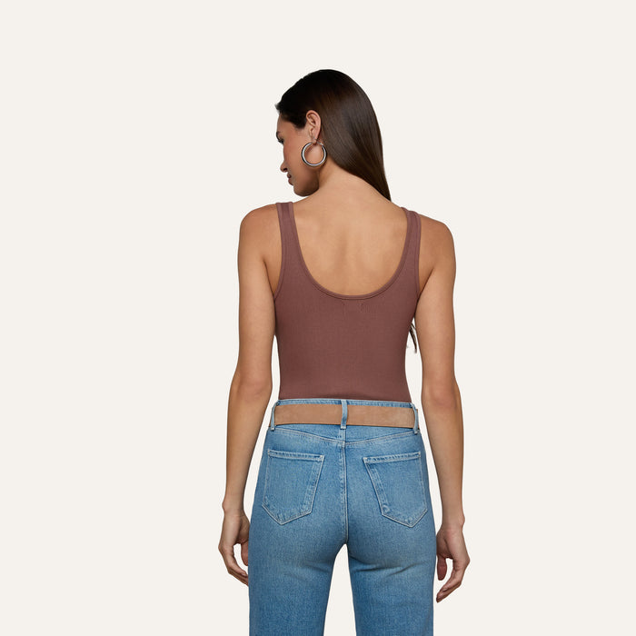 Woman facing away, wearing the Nica Scoopneck Bodysuit—a high stretch rib sleeveless brown top—paired with light blue high-waisted jeans with cutout details at the waist. She has long straight hair and large hoop earrings, standing against a plain light background.
