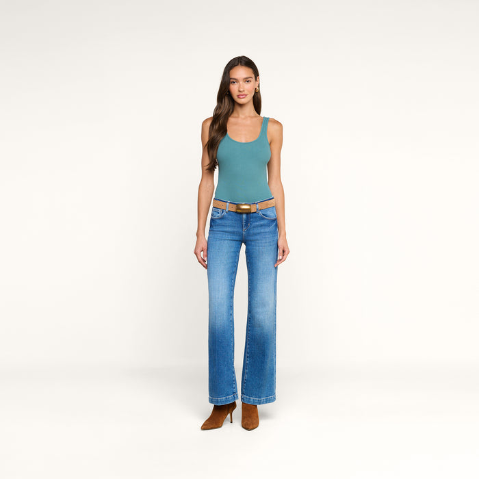 A woman stands against a plain white background wearing the Nica Scoopneck Bodysuit in teal, paired with blue flared jeans, a brown belt, and brown boots. Her long, wavy hair frames her face as she relaxes her arms at her sides.