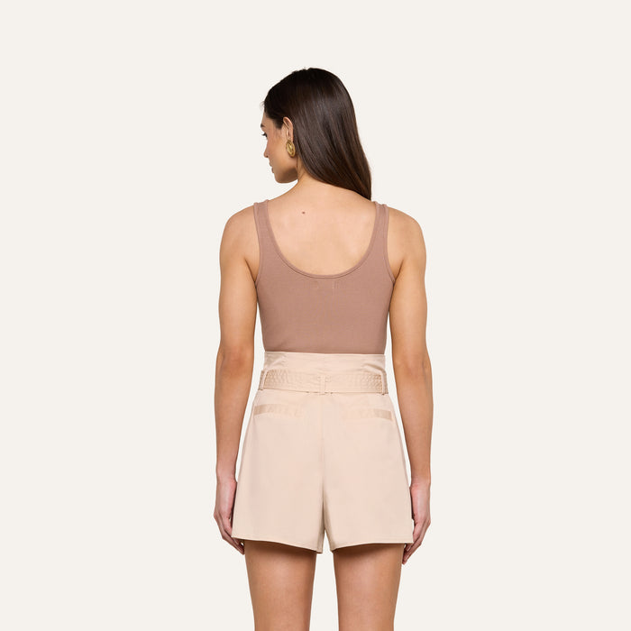A woman with long brown hair, wearing the Nica Scoopneck Bodysuit—a sleeveless taupe top with a sculpted fit—and high-waisted beige shorts stands facing away from the camera against a plain light background.