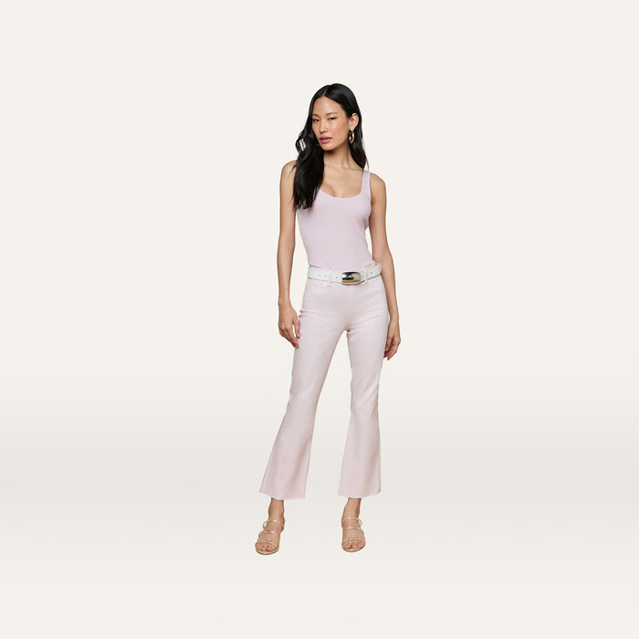 A woman stands against a plain background, wearing a light pink Nica Scoopneck Bodysuit by L’AGENCE, matching flared pants, a white belt, earrings, and nude heeled sandals.