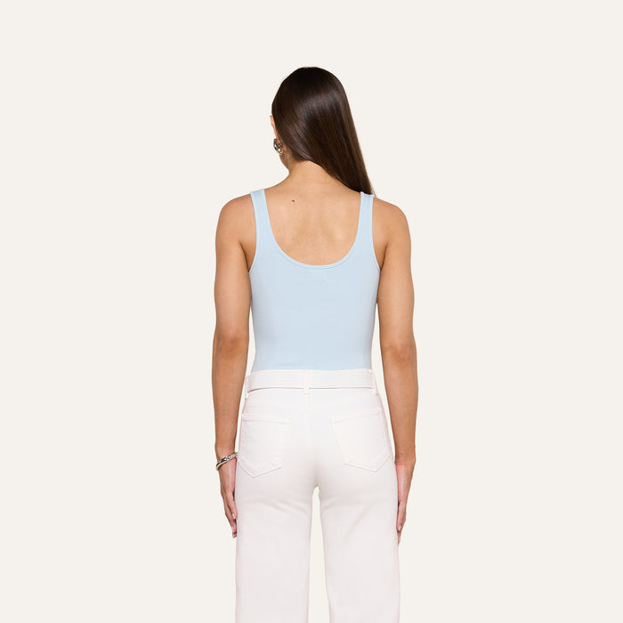 A woman with long brown hair, wearing the Nica Scoopneck Bodysuit in light blue and high-waisted white pants, stands facing away from the camera against a plain light background. The sculpted fit of the bodysuit highlights her silhouette.
