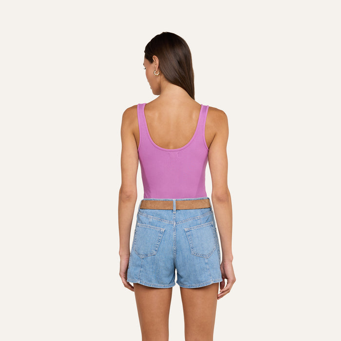 A woman stands with her back to the camera against a plain light background, wearing the Nica Scoopneck Bodysuit—a sleeveless purple tank top crafted from high recovery stretch rib—paired with light blue denim shorts.