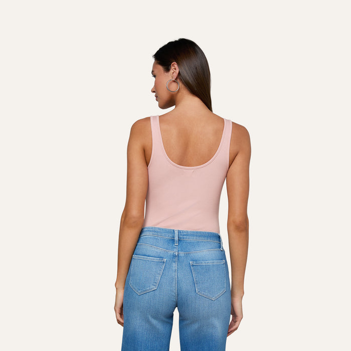 Woman with long brown hair and large hoop earrings, wearing the Nica Scoopneck Bodysuit in light pink high stretch rib and blue high-waisted jeans, shown from the back against an off-white background.