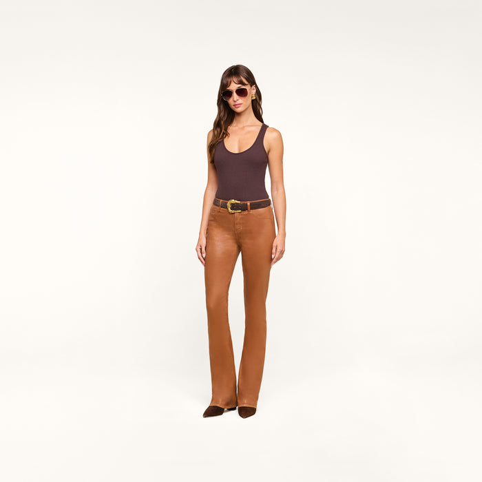A woman wearing sunglasses, a dark brown Nica Scoopneck Bodysuit, brown leather pants with a belt, and brown shoes stands against a plain white background.