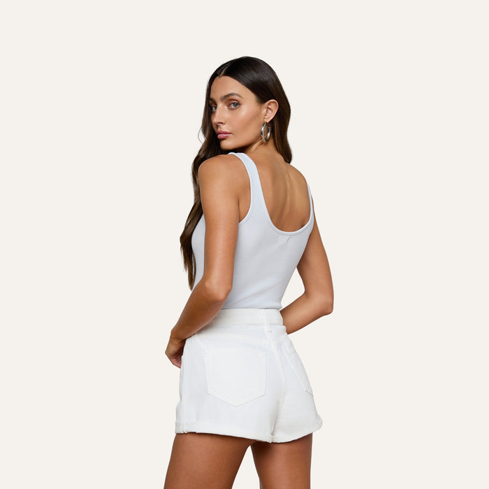 A woman with long brown hair wears the Nica Scoopneck Bodysuit in light blue and white high-waisted shorts, standing with her back slightly turned and looking over her shoulder against a plain light background.