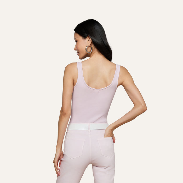A woman with long dark hair, wearing the Nica Scoopneck Bodysuit in light pink and matching pants, stands with her back to the camera, hand on her hip, against a plain light background.