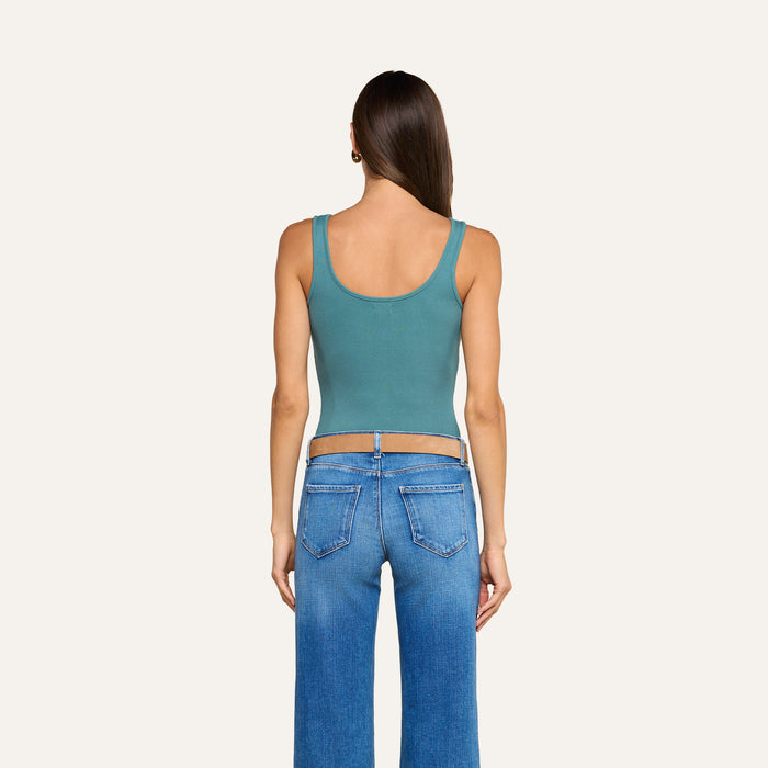 A woman with long brown hair stands facing away, wearing a teal Nica Scoopneck Bodysuit made from high recovery stretch rib and blue jeans. The background is plain and off-white.