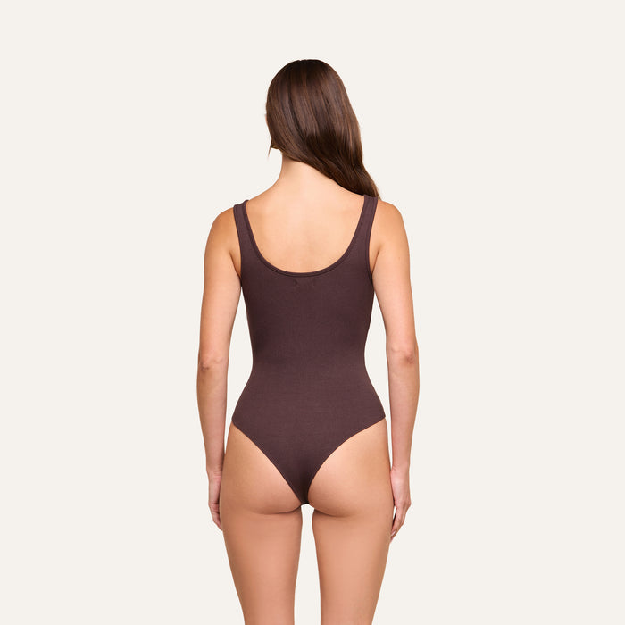 A woman with long brown hair stands facing away, wearing the Nica Scoopneck Bodysuit in dark brown stretch rib fabric against a plain light background.