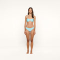 A woman stands facing forward against a plain background, wearing an ice blue Nicole Bikini Bottom made from stretch nylon and hoop earrings.
