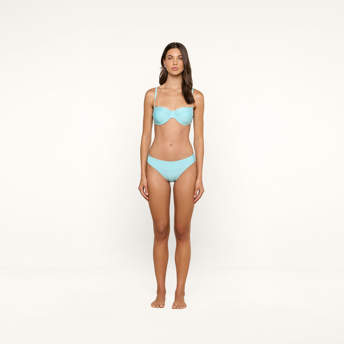 A woman stands facing forward against a plain background, wearing an ice blue Nicole Bikini Bottom made from stretch nylon and hoop earrings.