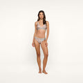 A woman stands barefoot against a plain white background, wearing the Nicole Bikini Bottom in a matching paisley print set with a bralette top. The low-rise bottoms are crafted from stretch nylon as she faces forward with a neutral expression.
