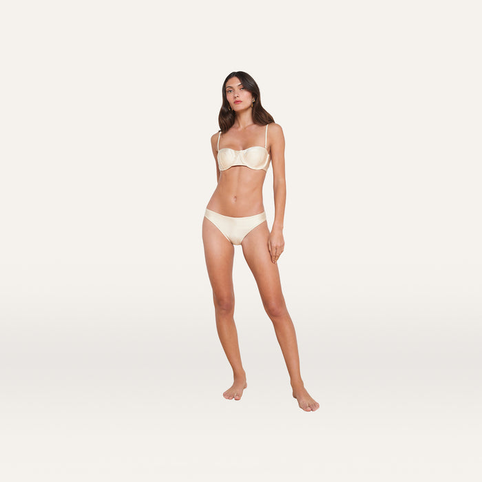 A woman stands barefoot against a plain white background, wearing a matching light-colored bra and the Nicole Bikini Bottom, crafted from soft fabric with four-way stretch for moderate coverage.