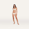 A woman stands barefoot against a plain white background, wearing a matching light-colored bra and the Nicole Bikini Bottom, crafted from soft fabric with four-way stretch for moderate coverage.