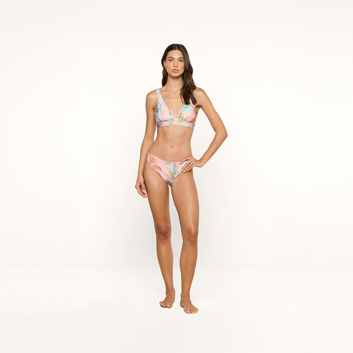 A woman stands barefoot against a plain white background, wearing the Nicole Bikini Bottom in a pastel floral pattern crafted from stretch nylon, with one hand resting on her hip and a neutral expression on her face.