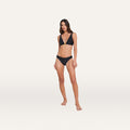 Woman standing against a plain white background, wearing the Nicole Bikini Bottom in black with moderate coverage, paired with a matching triangle bikini top, both made from four-way stretch nylon, looking forward with a neutral expression.