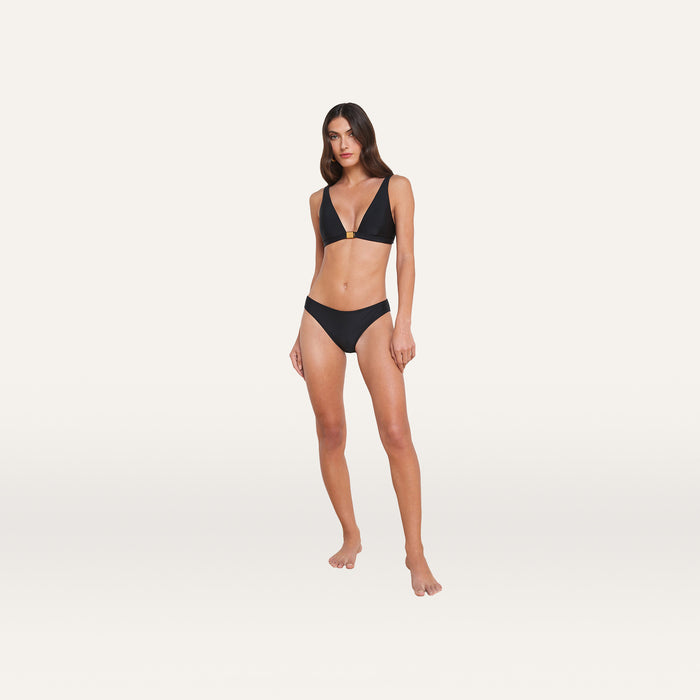 Woman standing against a plain white background, wearing the Nicole Bikini Bottom in black with moderate coverage, paired with a matching triangle bikini top, both made from four-way stretch nylon, looking forward with a neutral expression.