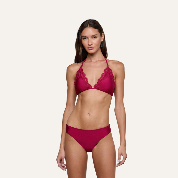 A woman stands facing forward, wearing the Nicole Bikini Bottom in black cherry with lace trim and a matching top. The background is plain and off-white.