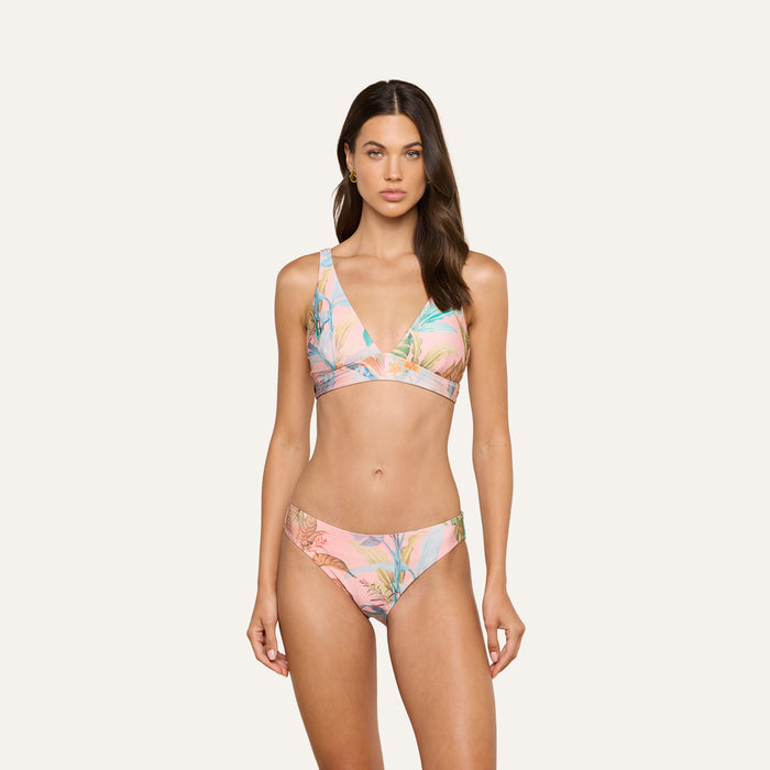 A woman stands against a plain background wearing the Nicole Bikini Bottom, a pink bikini featuring a tropical floral and palm leaf print. Crafted from stretch nylon for comfort and durability, the swimwear offers UPF 50 protection. She has long, dark hair, looks forward, and keeps her arms relaxed at her sides.