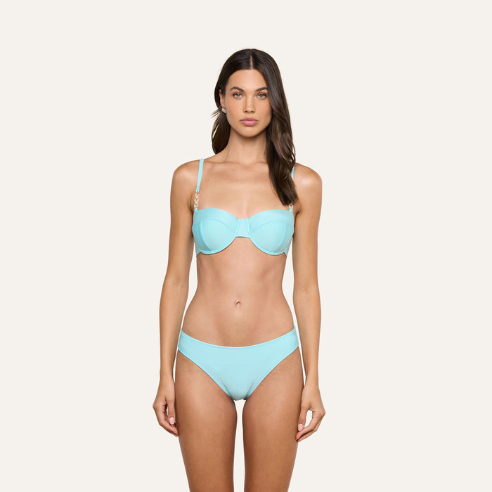 A woman is standing and facing forward, wearing the Nicole Bikini Bottom in ice blue with gold accents on the straps, made from stretch nylon, against a plain light background.