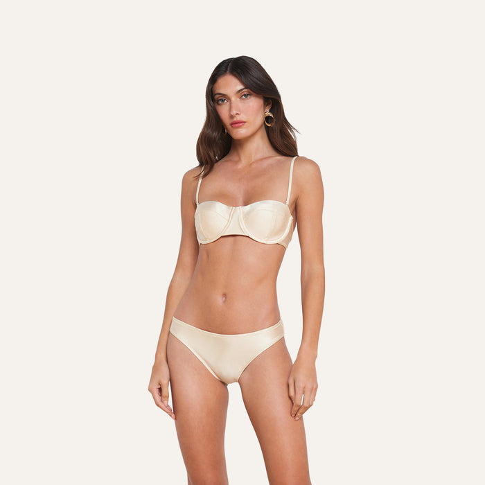A woman stands against a plain background wearing a matching cream-colored bra and the Nicole Bikini Bottom, which offers moderate coverage. She has long brown hair and is looking slightly to the side with a neutral expression.