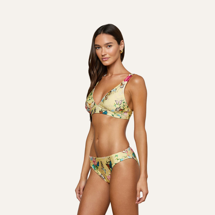 A woman poses in the Nicole Bikini Bottom, featuring a yellow base with a colorful floral pattern and moderate coverage, against a plain light background.