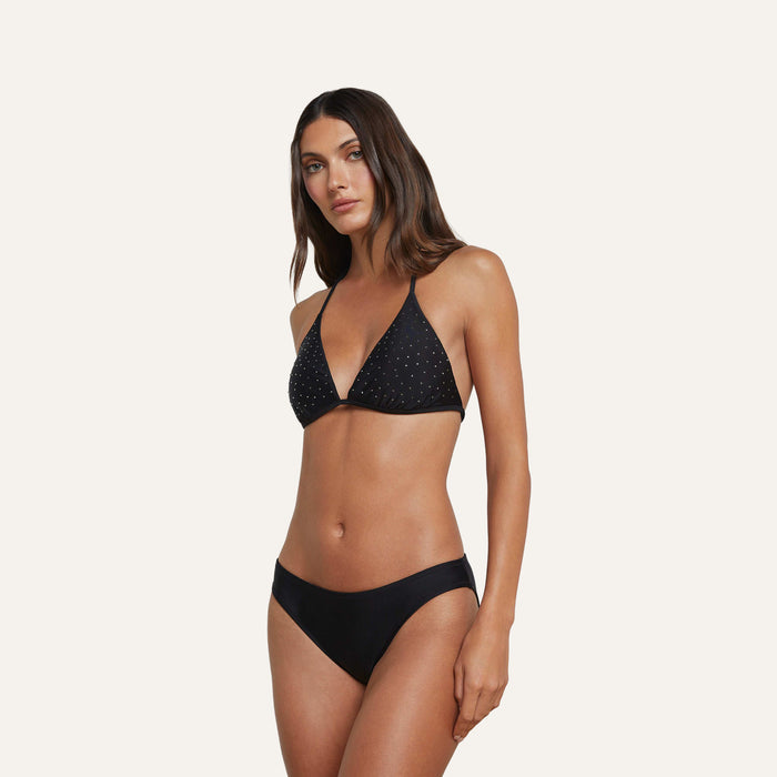 Woman wearing a versatile black bikini with a studded or dotted pattern on the top and moderate coverage Nicole Bikini Bottom, standing against a plain light background.