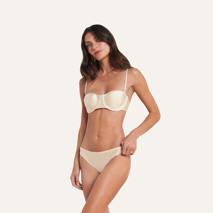 A woman stands against a plain background, wearing a matching cream-colored lingerie set featuring a balconette bra and the Nicole Bikini Bottom with moderate coverage. She looks toward the camera with a neutral expression.